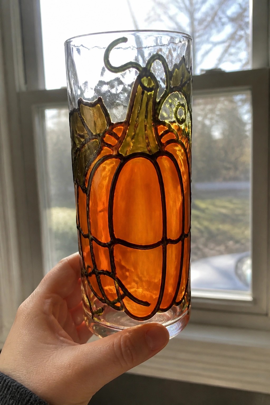Hand holding tall glass with orange pumpkin and green leaf stained-glass design