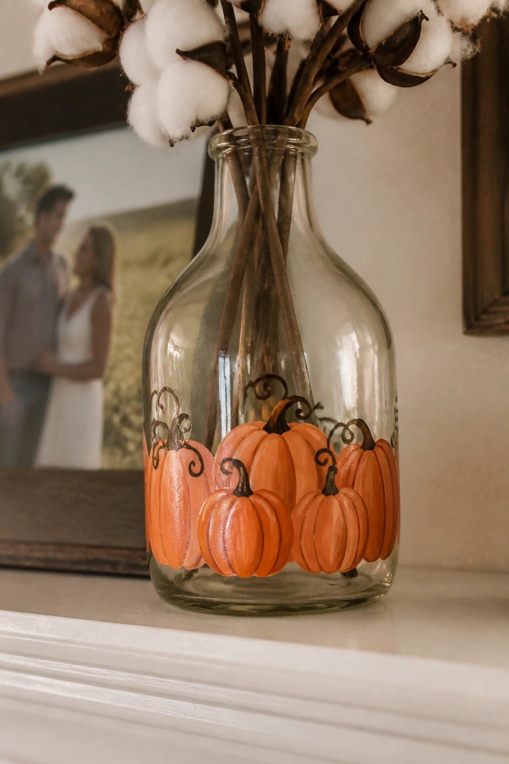 A clear glass bottle painted with orange pumpkins and black stems holds cotton branches on a white surface.