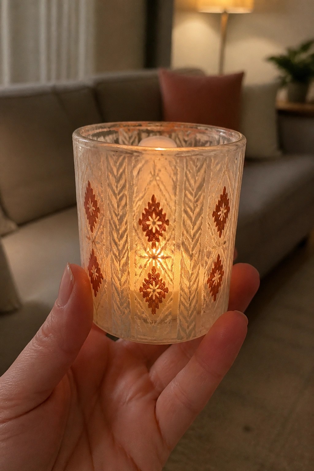 Hand holding lit candle in etched glass holder with red diamond patterns.