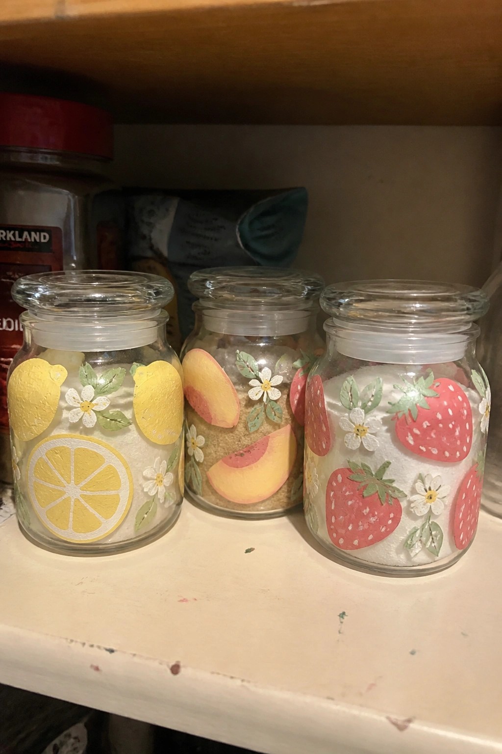 Three glass jars with lemon, peach, and strawberry stickers holding sugar on shelf