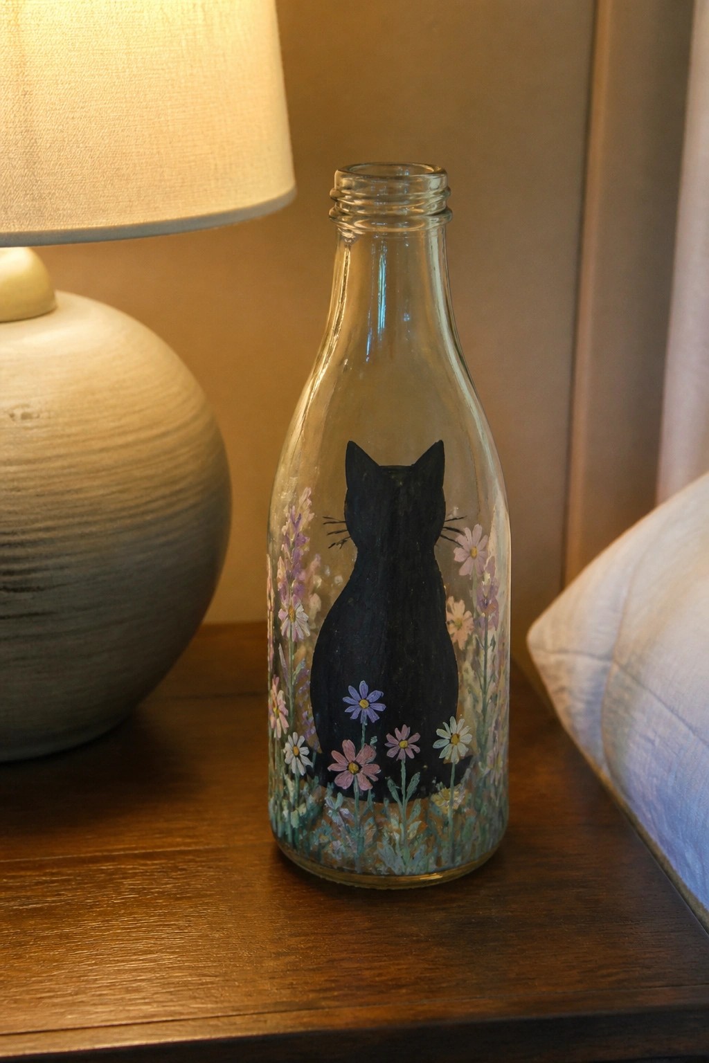 Glass bottle painted with black cat silhouette amid colorful wildflowers on wooden table