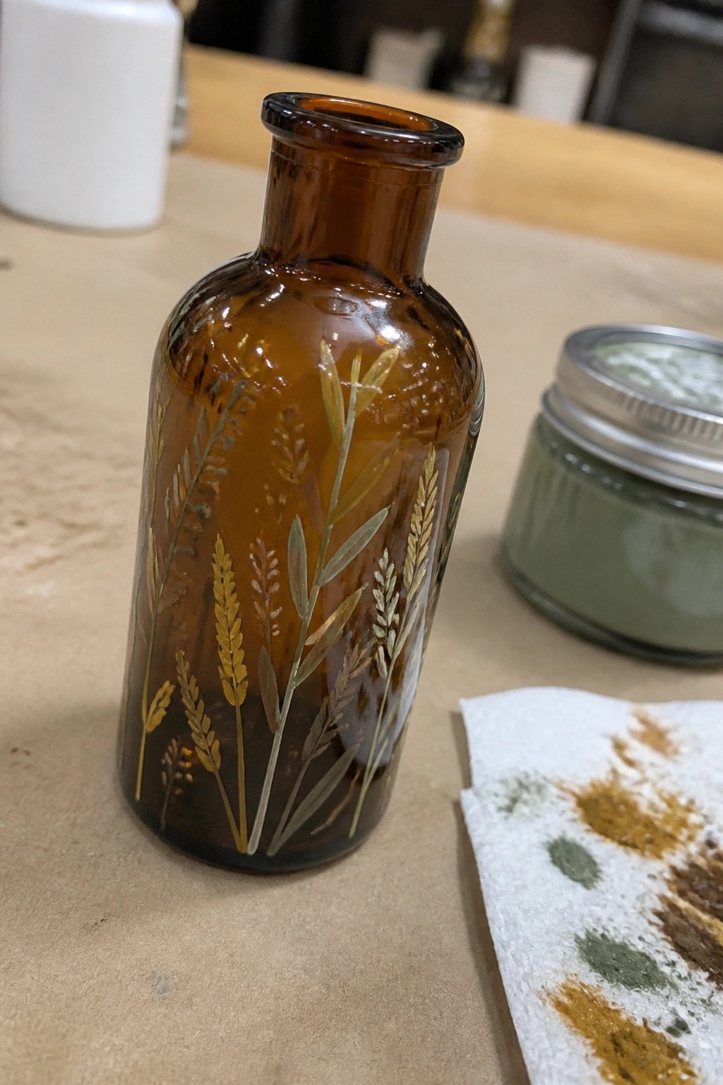 Amber glass bottle with golden wheat designs beside green jar and paint-stained towel