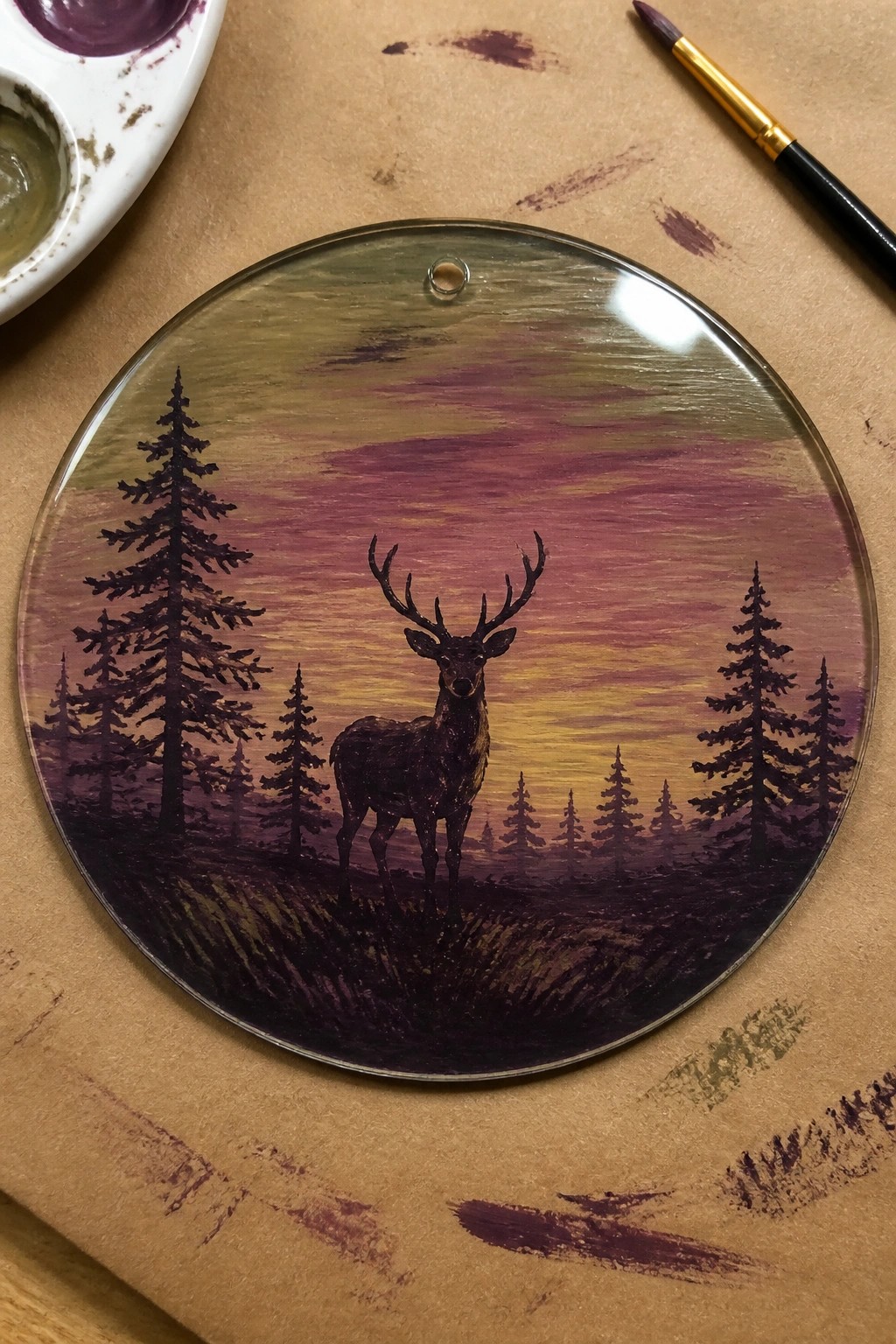 Circular glass deer painting amid pine trees under purple-yellow sunset, with palette and brush nearby.