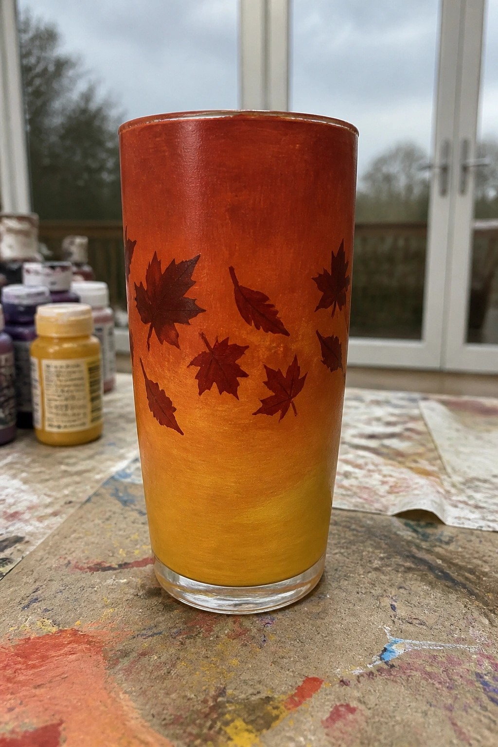 Orange-yellow gradient glass tumbler with dark maple leaves on paint-splattered table