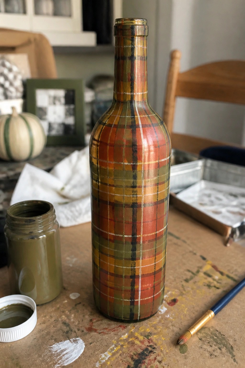 Plaid-painted bottle in autumn hues on paint-splattered wooden table with supplies.