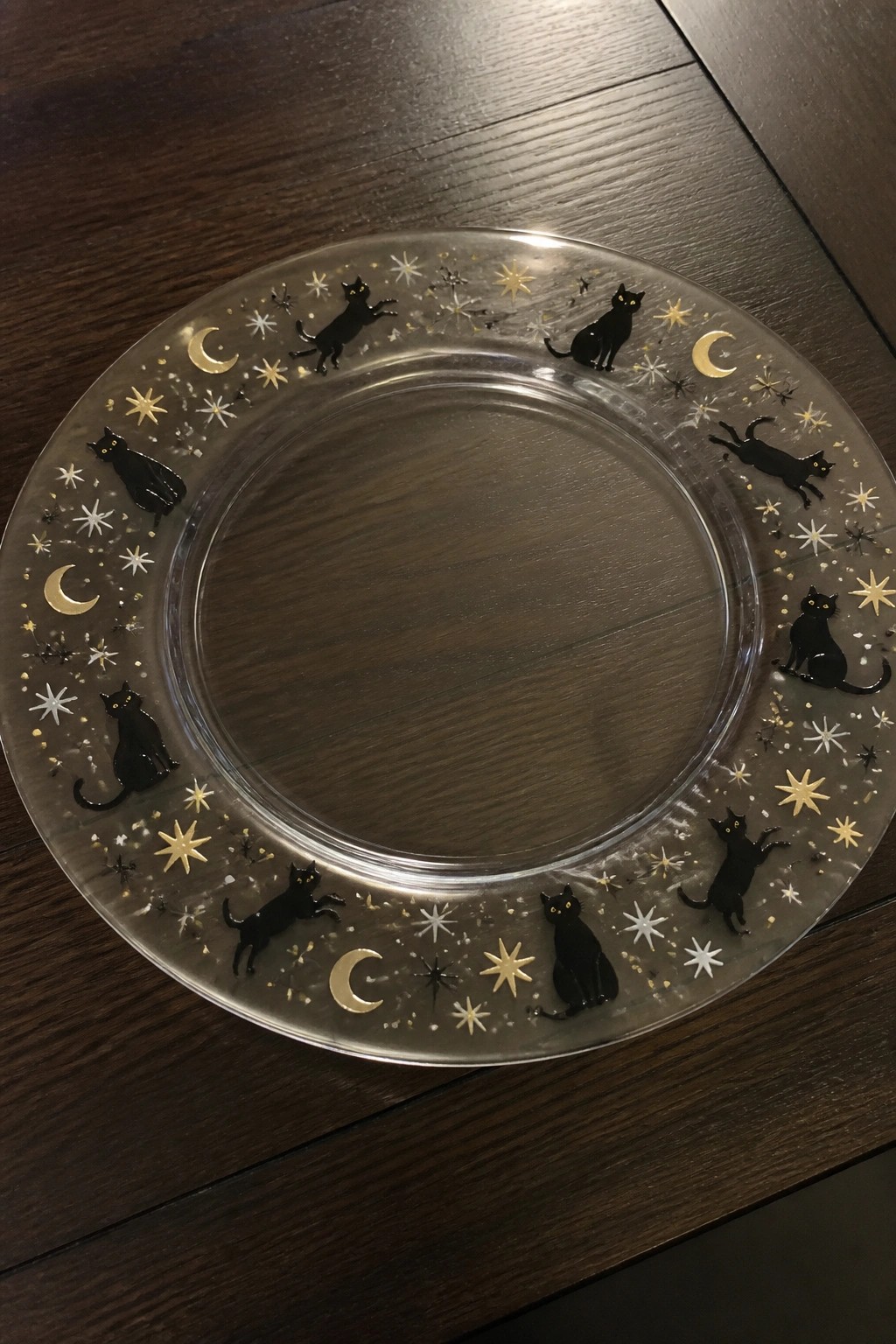 A clear round glass plate with black cat silhouettes, gold moons, and stars painted around the rim on a wooden surface.