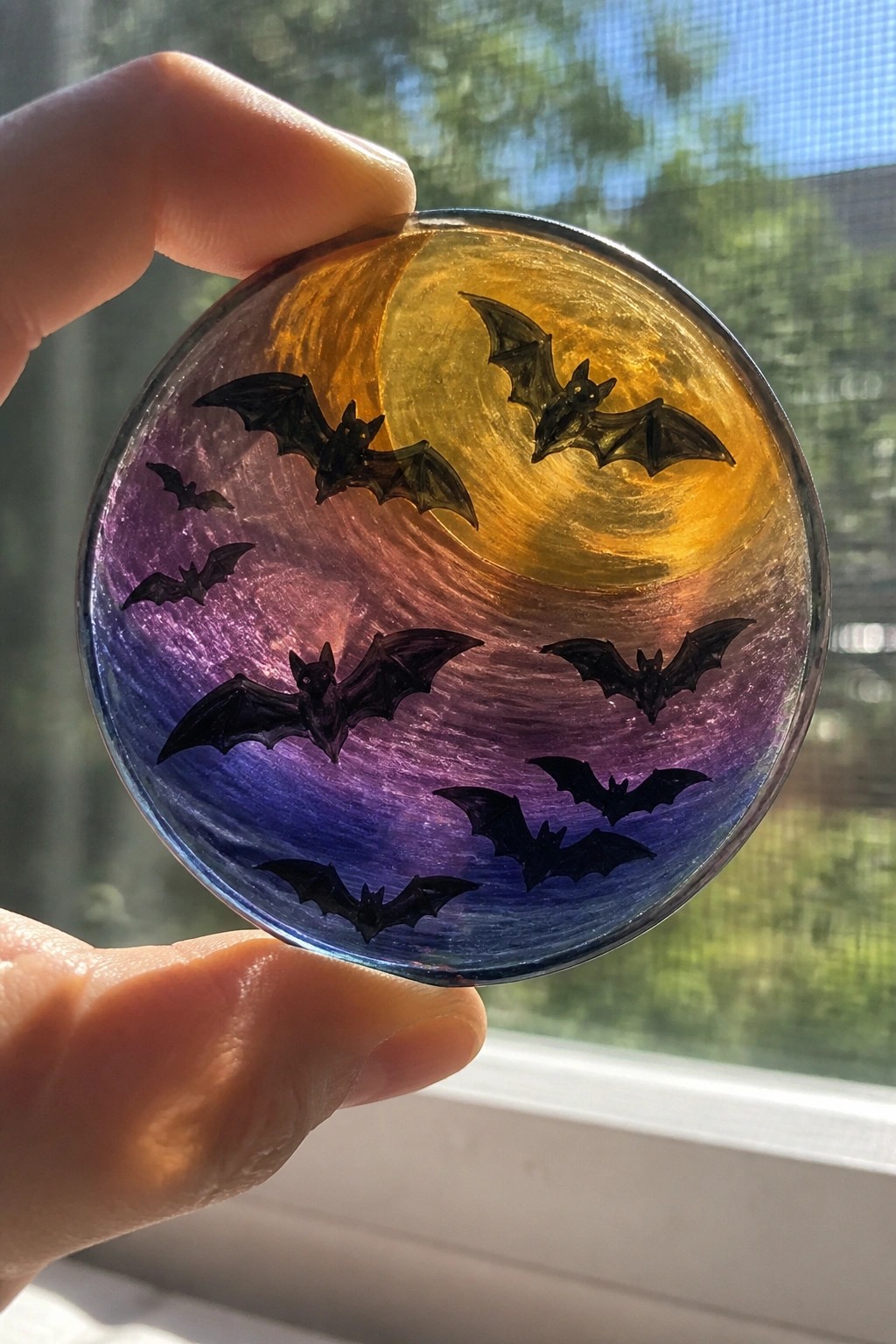 Hand holding round resin disc with black bats on purple-orange gradient swirls