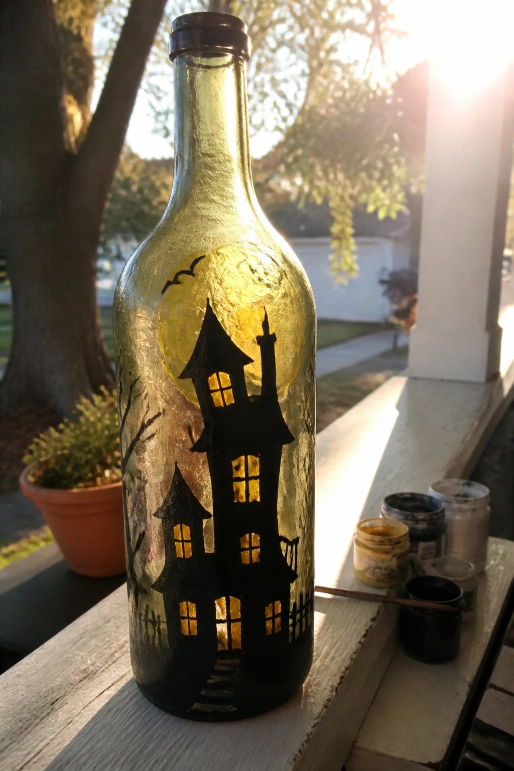Illuminated glass bottle with black haunted house, moon, bats and paint jars nearby