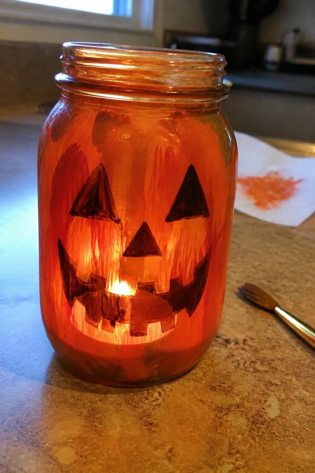 Illuminated orange mason jar with painted jack-o'-lantern face glowing on kitchen counter