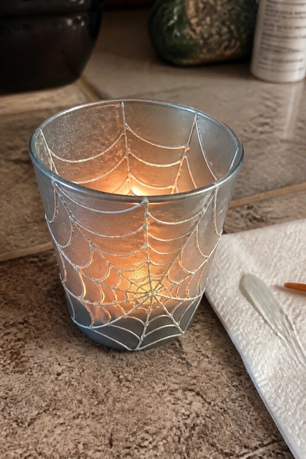 Lit glass votive with silver spiderweb design glowing on speckled countertop.