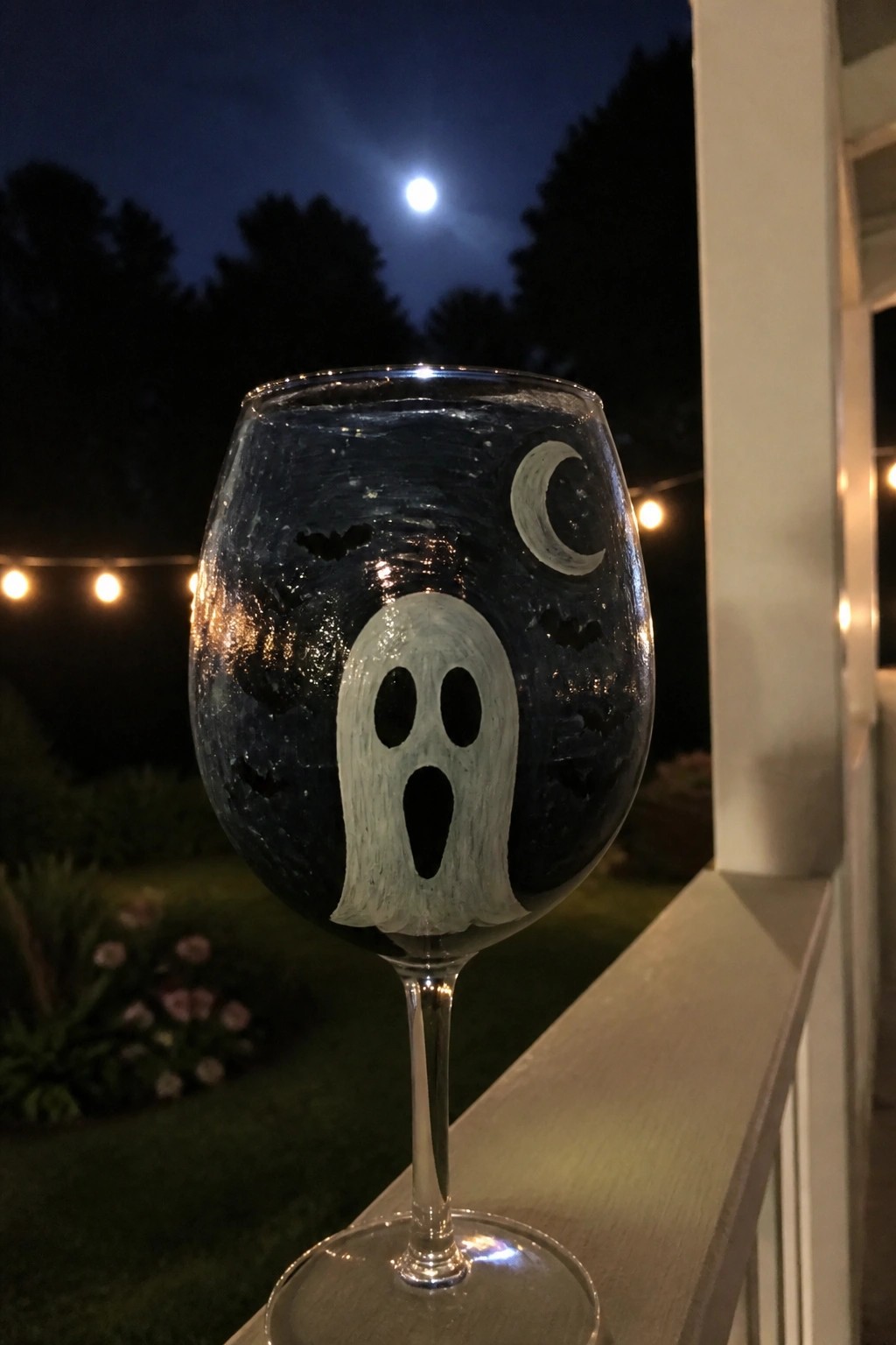Painted wine glass with ghost, bats, and moon on railing under night sky