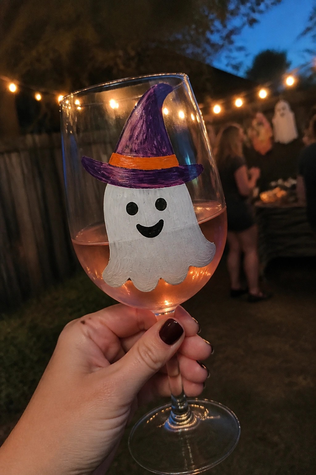 Hand holding wine glass painted with smiling ghost in purple witch hat at night party