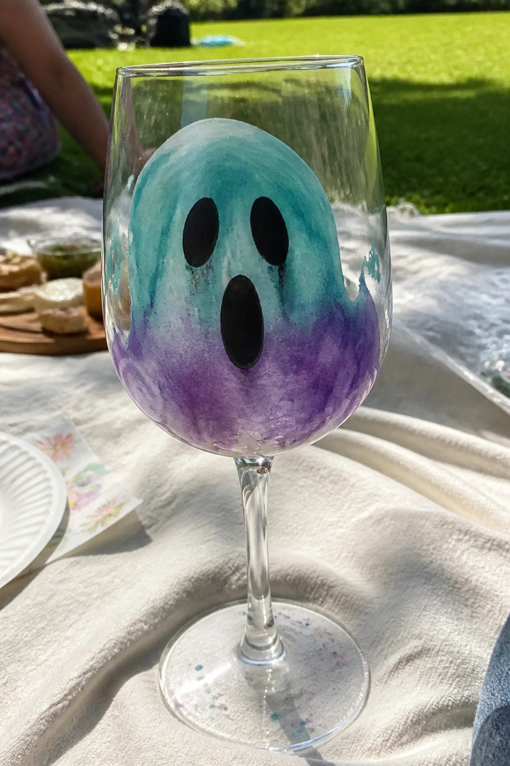 Wine glass with blue-purple watercolor ghost face on outdoor picnic blanket.