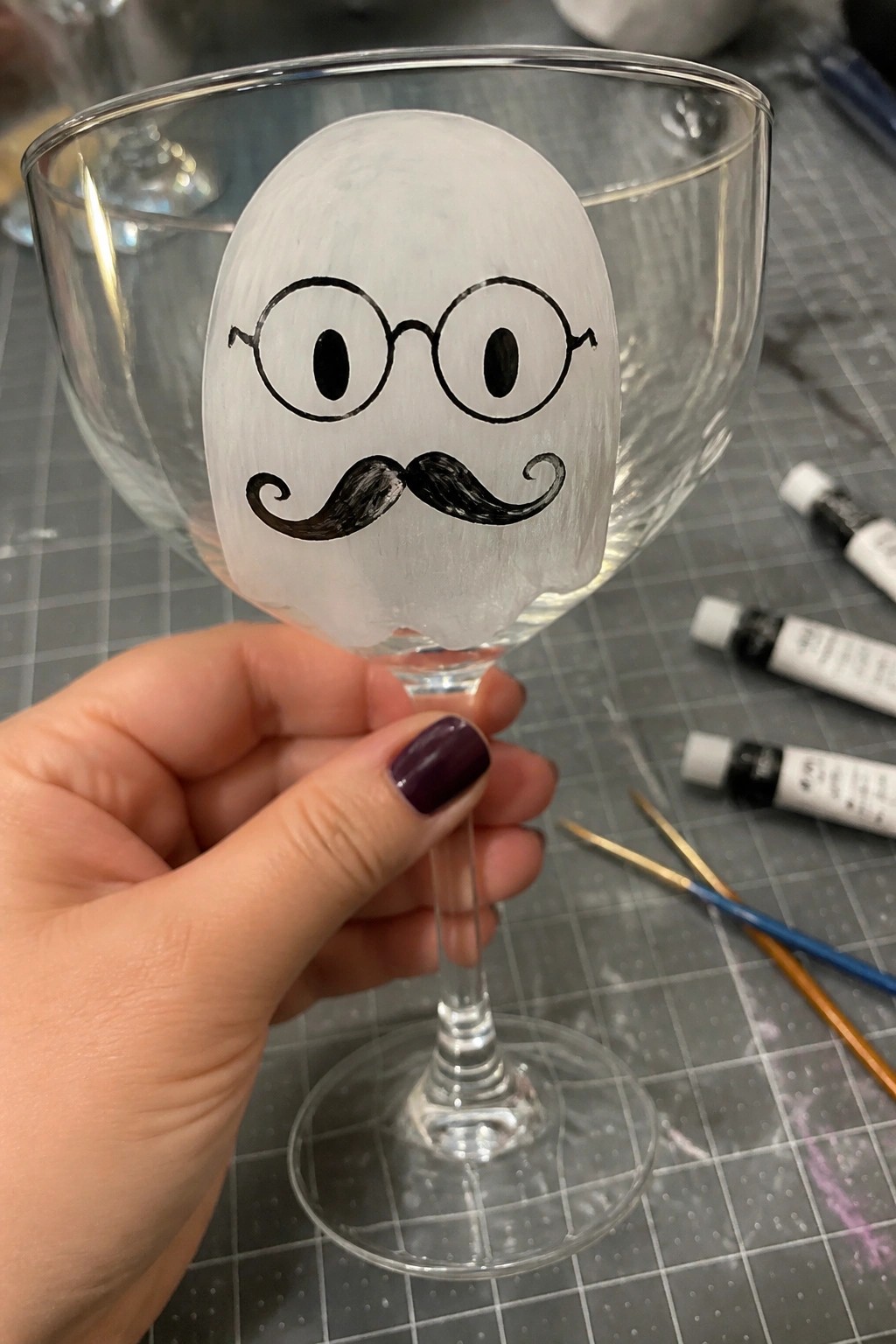 A hand holds a clear wine glass with a white painted ghost face that has round glasses and a black mustache.