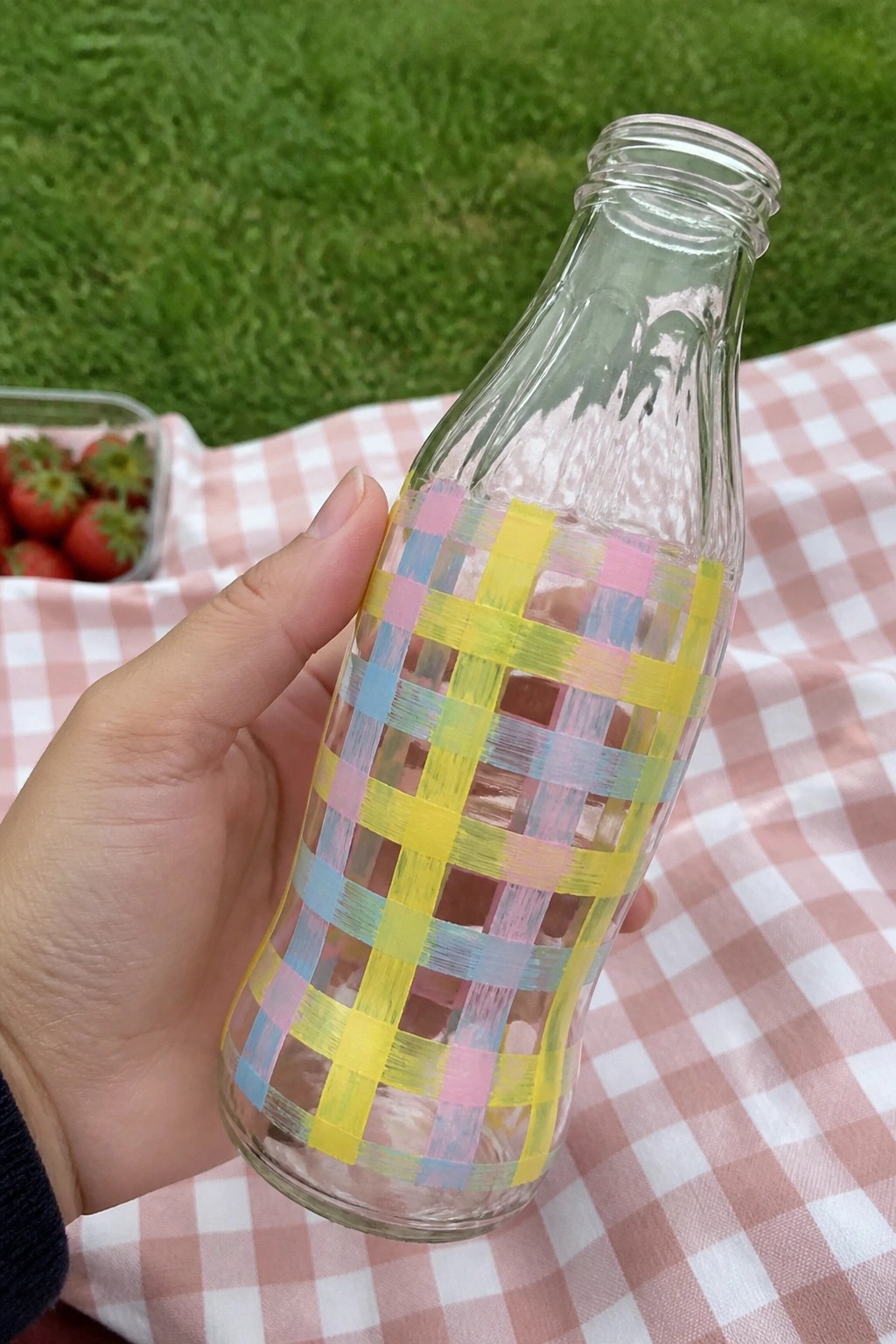 Hand holding glass bottle with yellow pink blue plaid tape on pink checkered picnic blanket