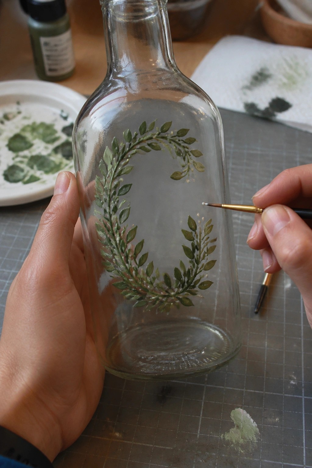 Hands painting green leaf wreath on clear glass bottle with fine brush