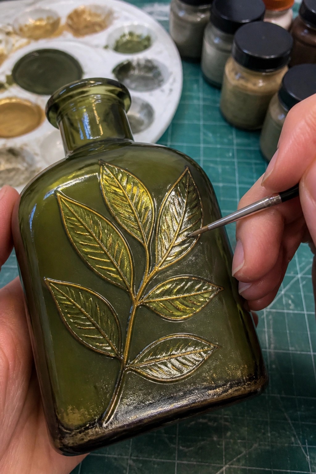 Hand applying gold paint with fine brush to embossed leaves on green glass bottle.
