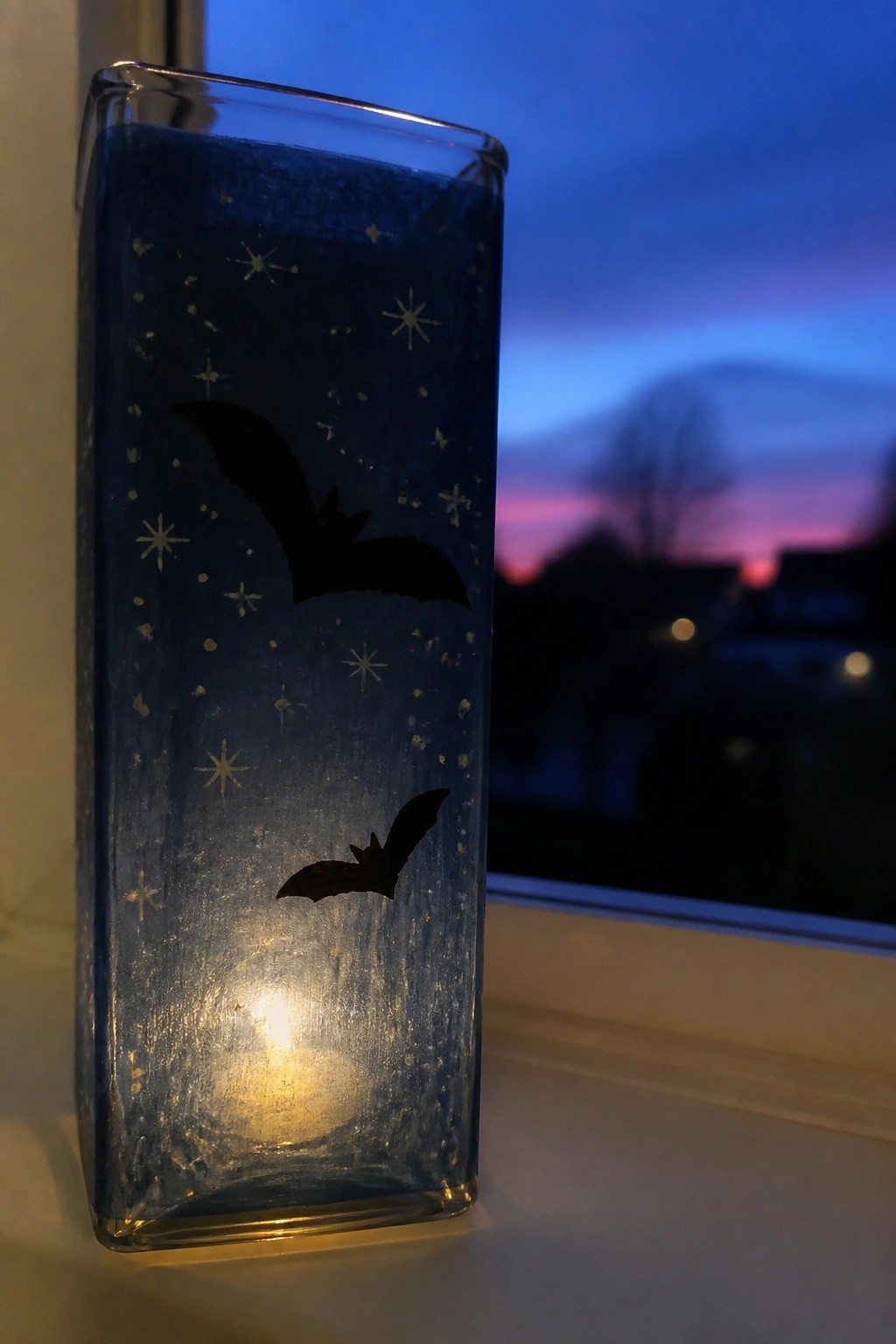 Lit blue glass candle holder with bats and stars, glowing on windowsill at dusk