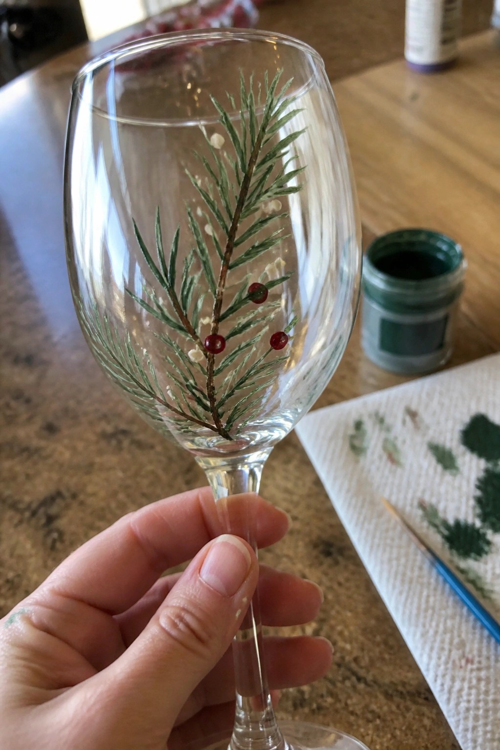 Hand holding wine glass painted with green pine branch, red berries on wooden table