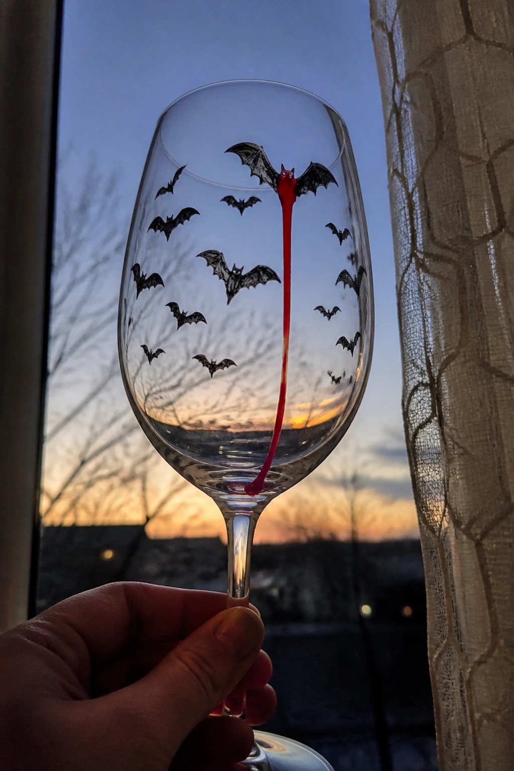 Hand holding wine glass with black bats and dripping red liquid at sunset