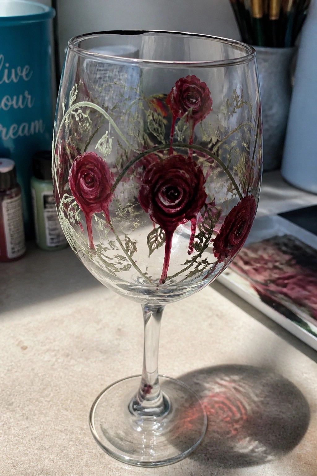 Hand-painted wine glass with dark red roses, green vines, and dripping paint accents