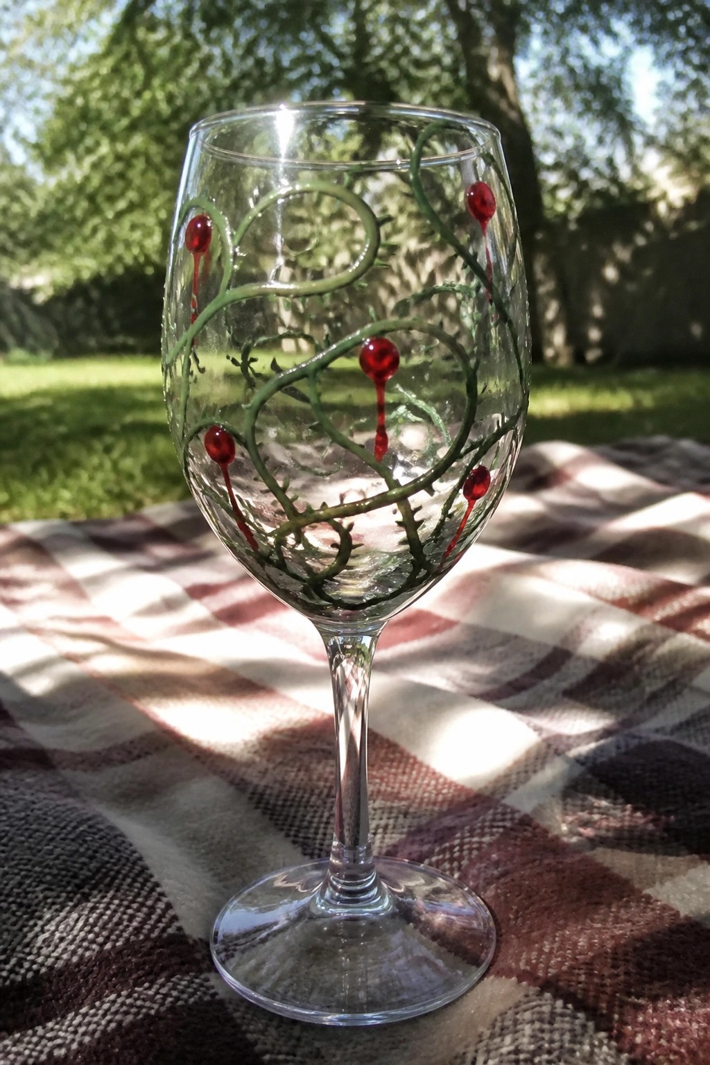 A clear wine glass with green thorny vines and red dripping accents rests on a plaid blanket outdoors.