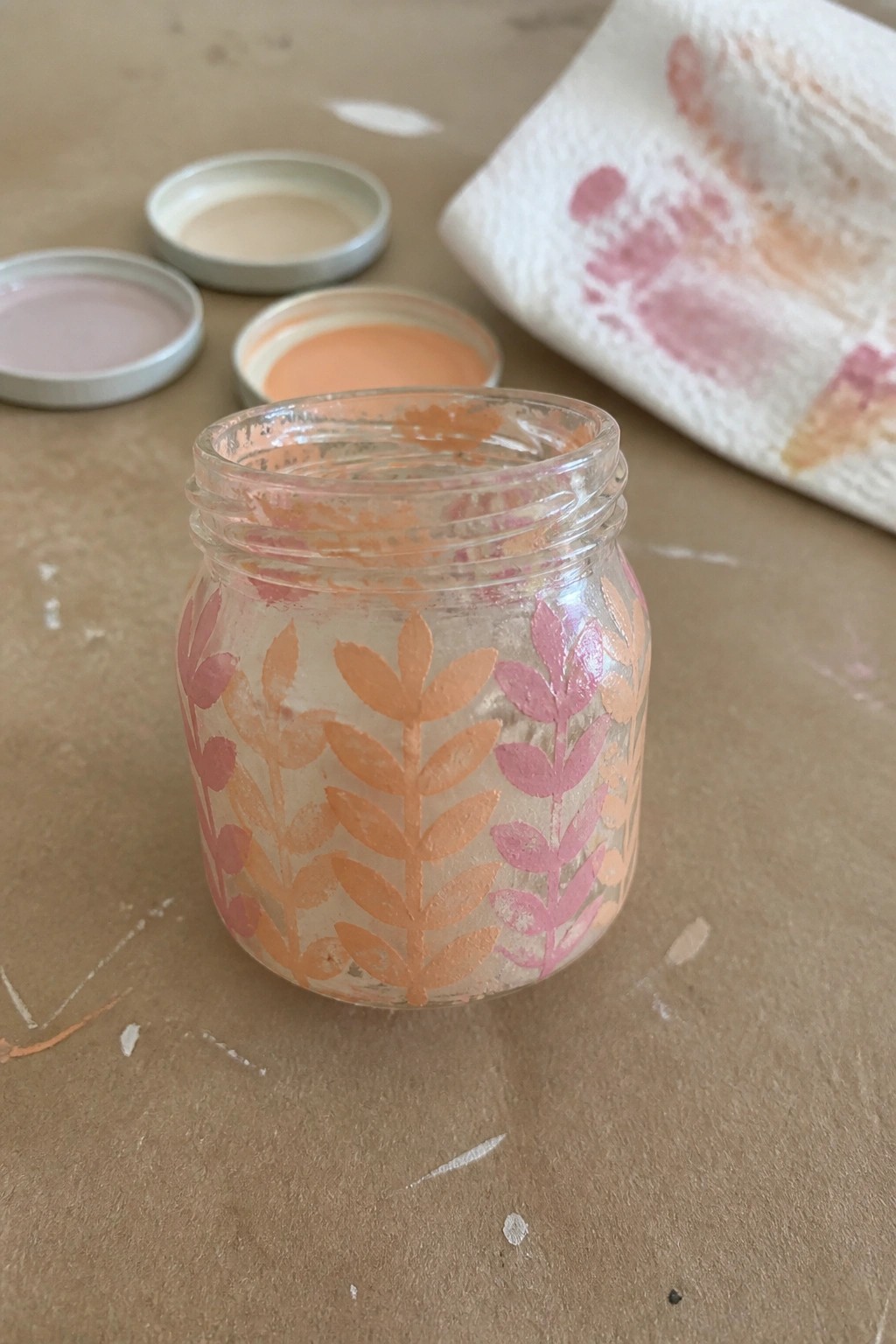 Glass jar painted with pink and orange leaves amid paint lids on brown surface