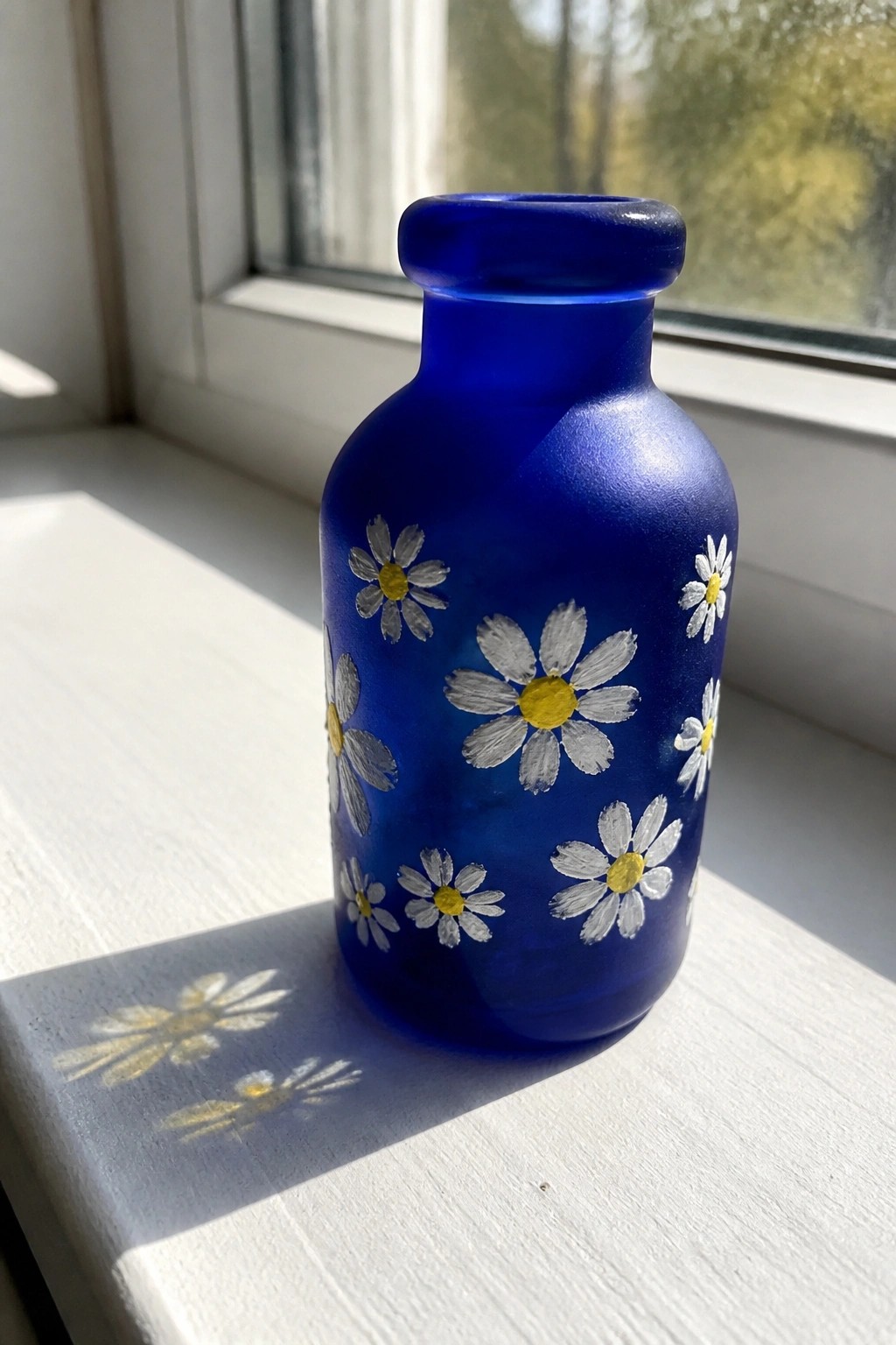 Blue glass bottle with painted white daisies on sunlit white windowsill