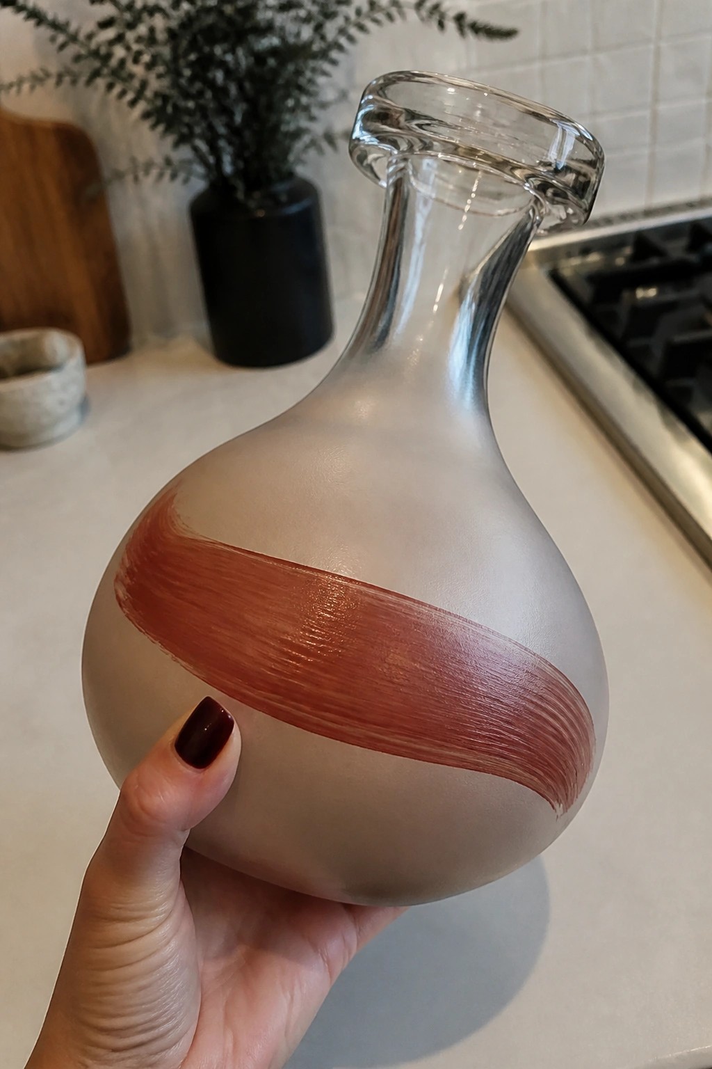 Hand with dark nails holds frosted glass carafe with terracotta stripe.