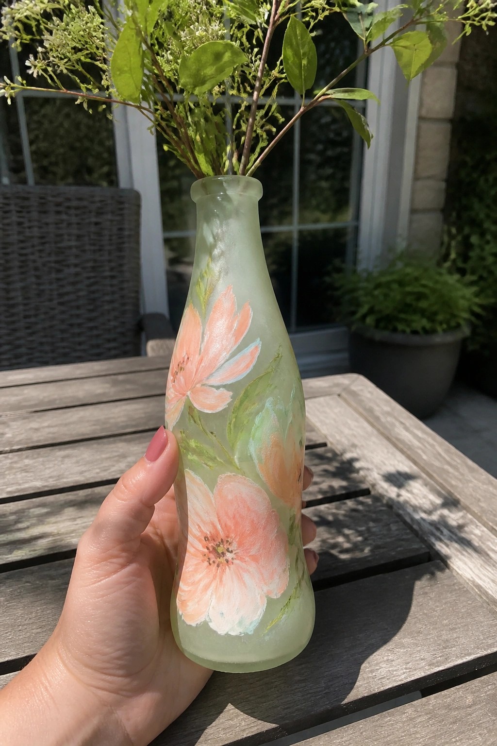 Hand holding frosted green bottle with pink floral paintings on wooden table outdoors.