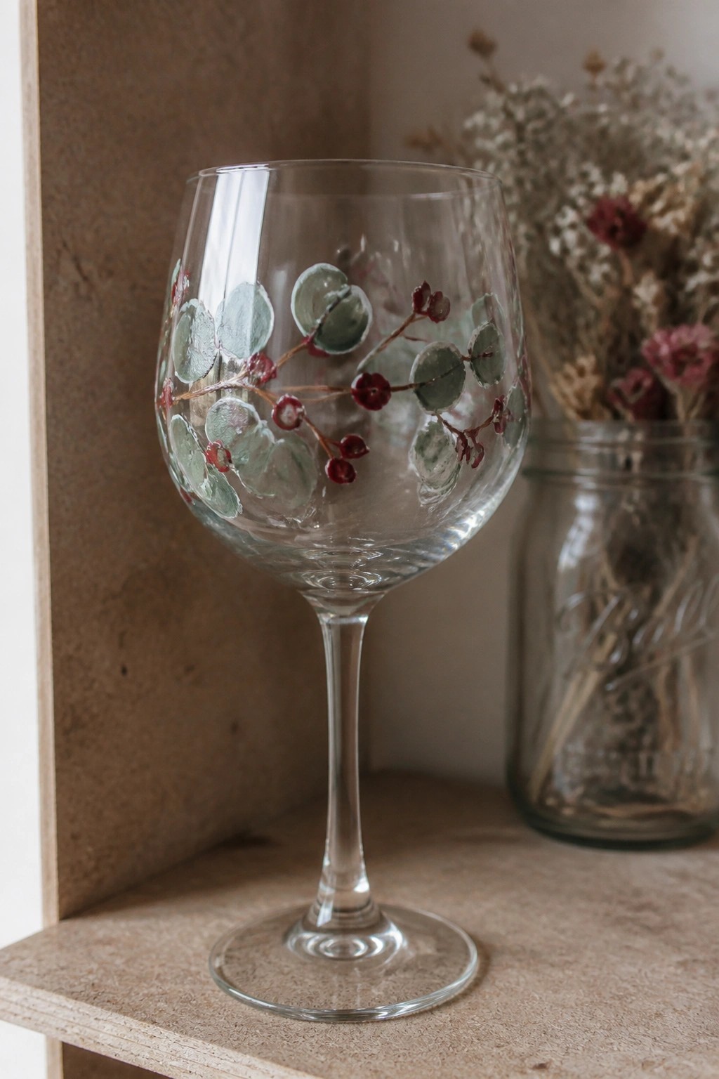 Hand-painted wine glass with green leaves and red berries on wooden shelf