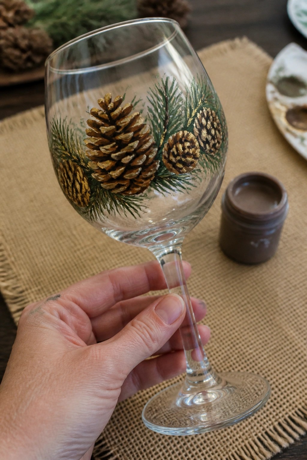 Hand holding wine glass painted with pinecones and green branches beside paint jar