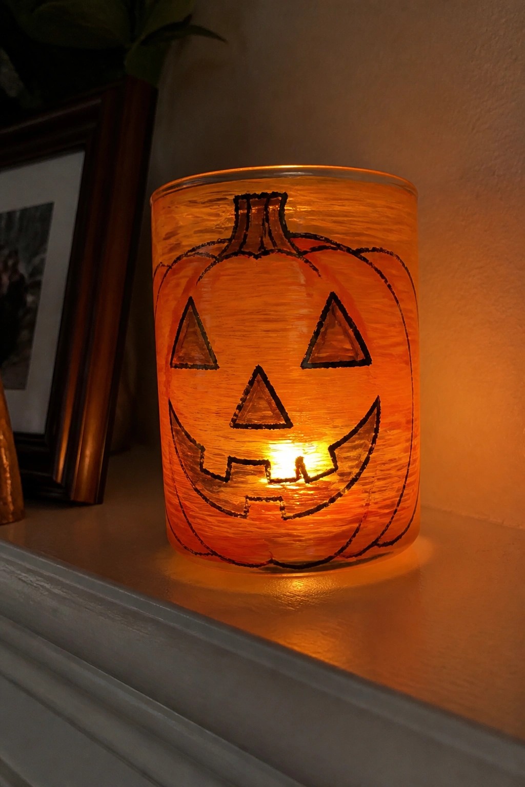 Glowing orange jack-o'-lantern candle holder on white mantel with frame nearby.