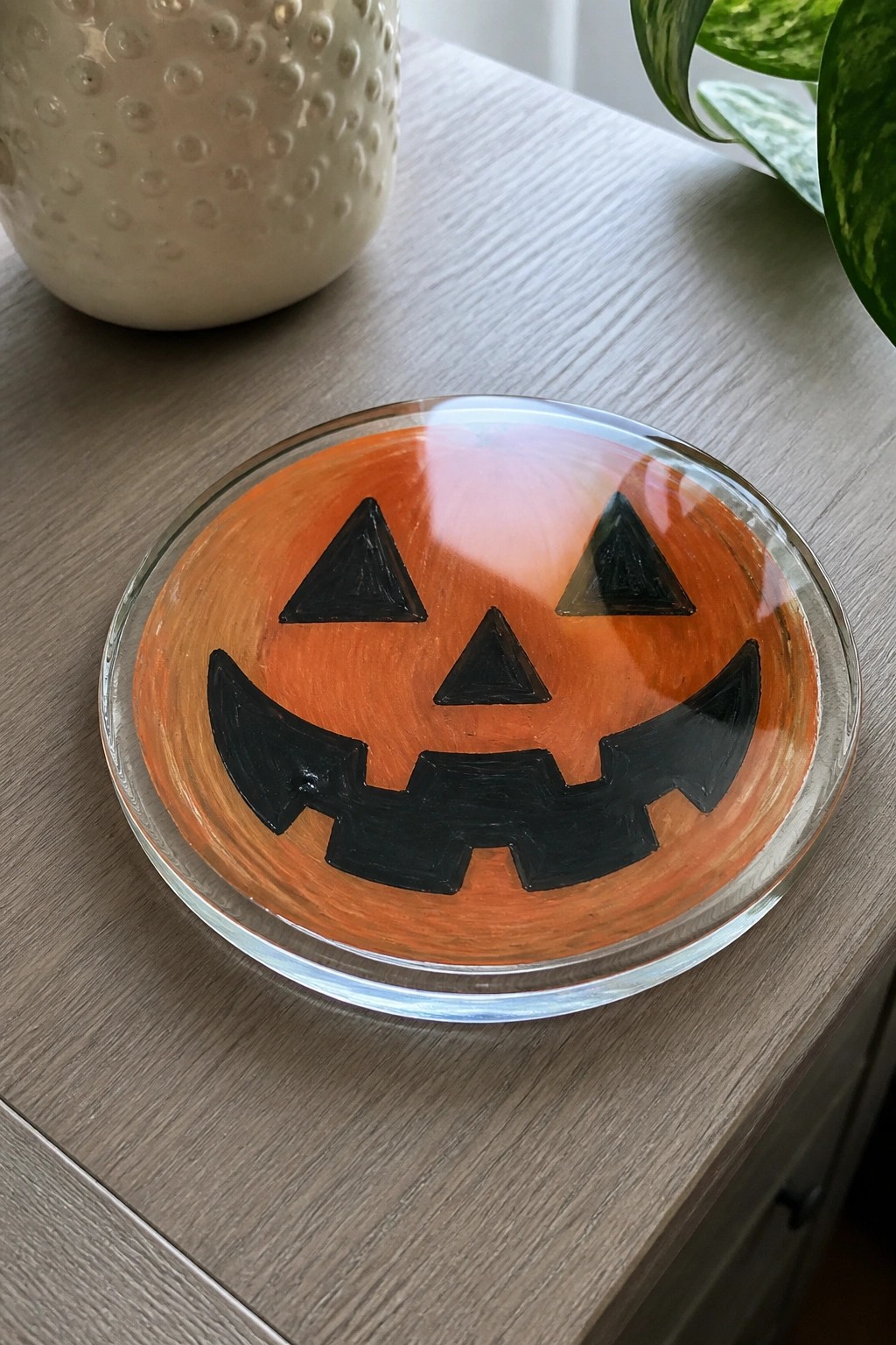 Jack-o'-lantern painted on round glass plate atop wooden table with plant nearby.
