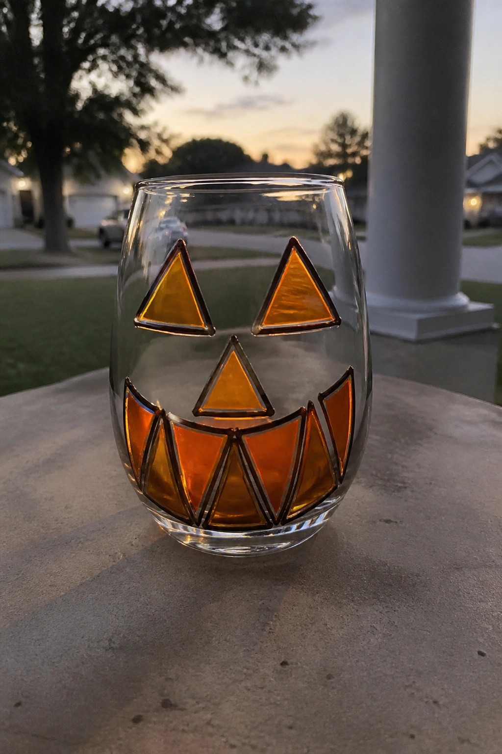 Stemless glass with orange stained-glass jack-o'-lantern face on concrete at dusk