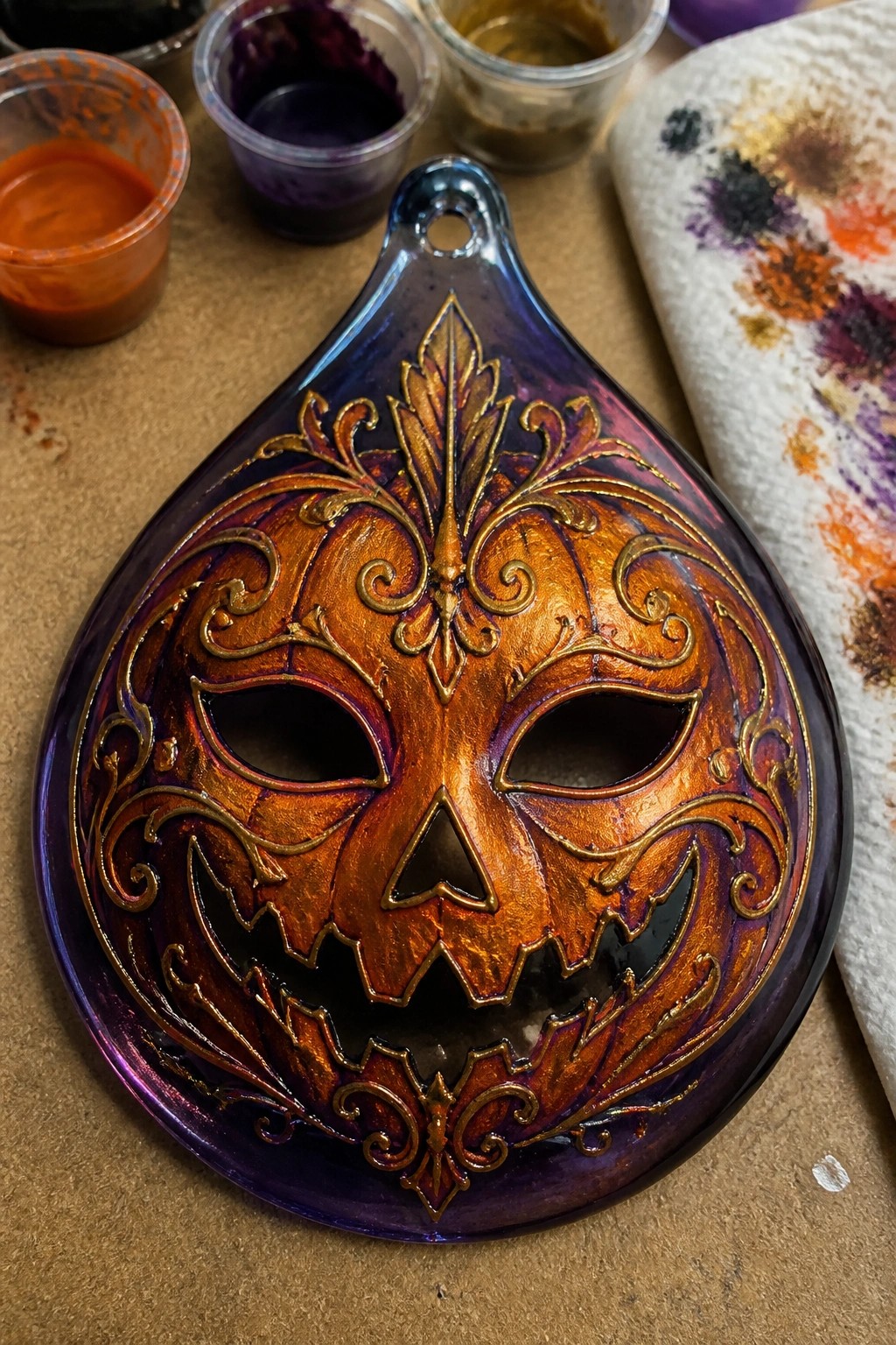 Ornate orange pumpkin mask with gold filigree on purple-blue amid paint cups