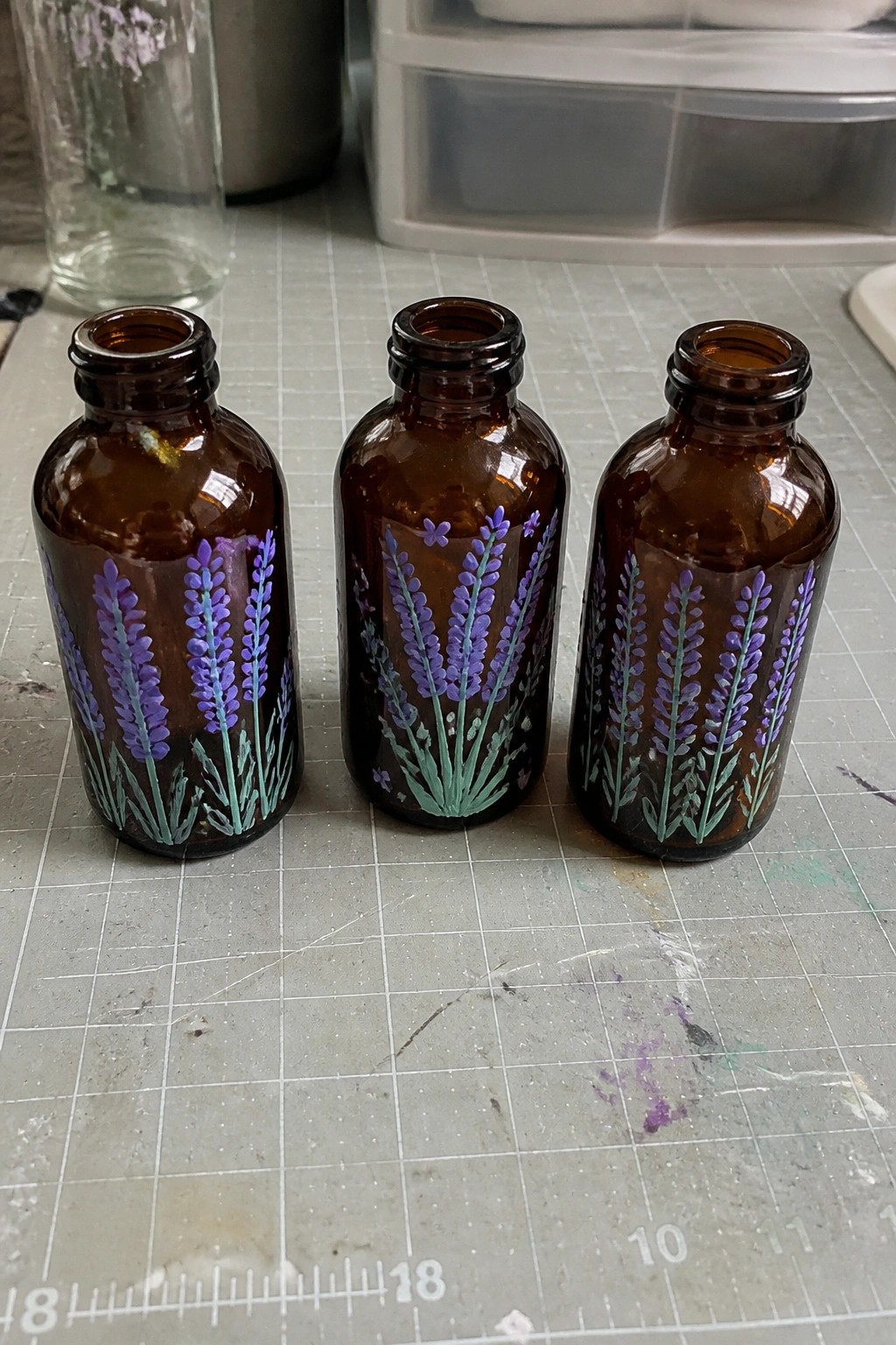 Three brown glass bottles with purple lavender paintings on a gray grid mat