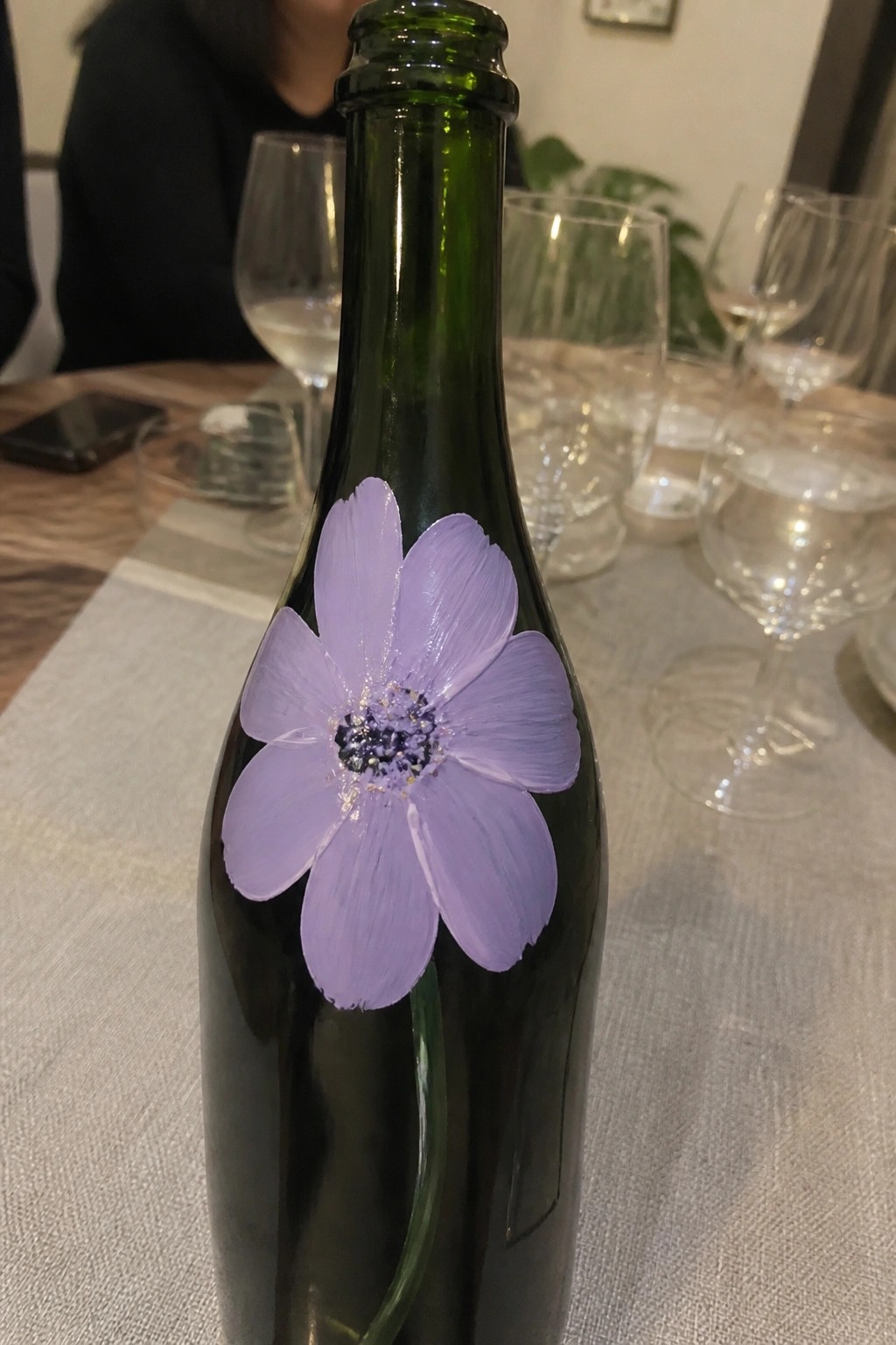 A dark green glass bottle with a large purple painted flower stands on a dining table surrounded by wine glasses.