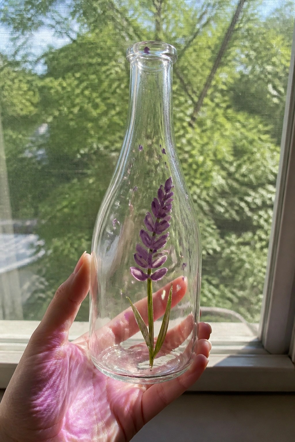 Hand holding clear glass bottle with purple lavender painting by sunlit window