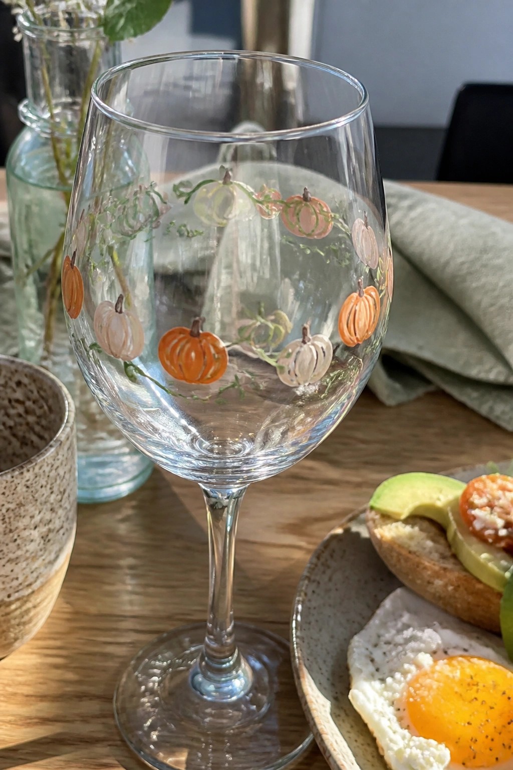 A clear wine glass painted with small orange, peach, and white pumpkins connected by green vines on a wooden table.