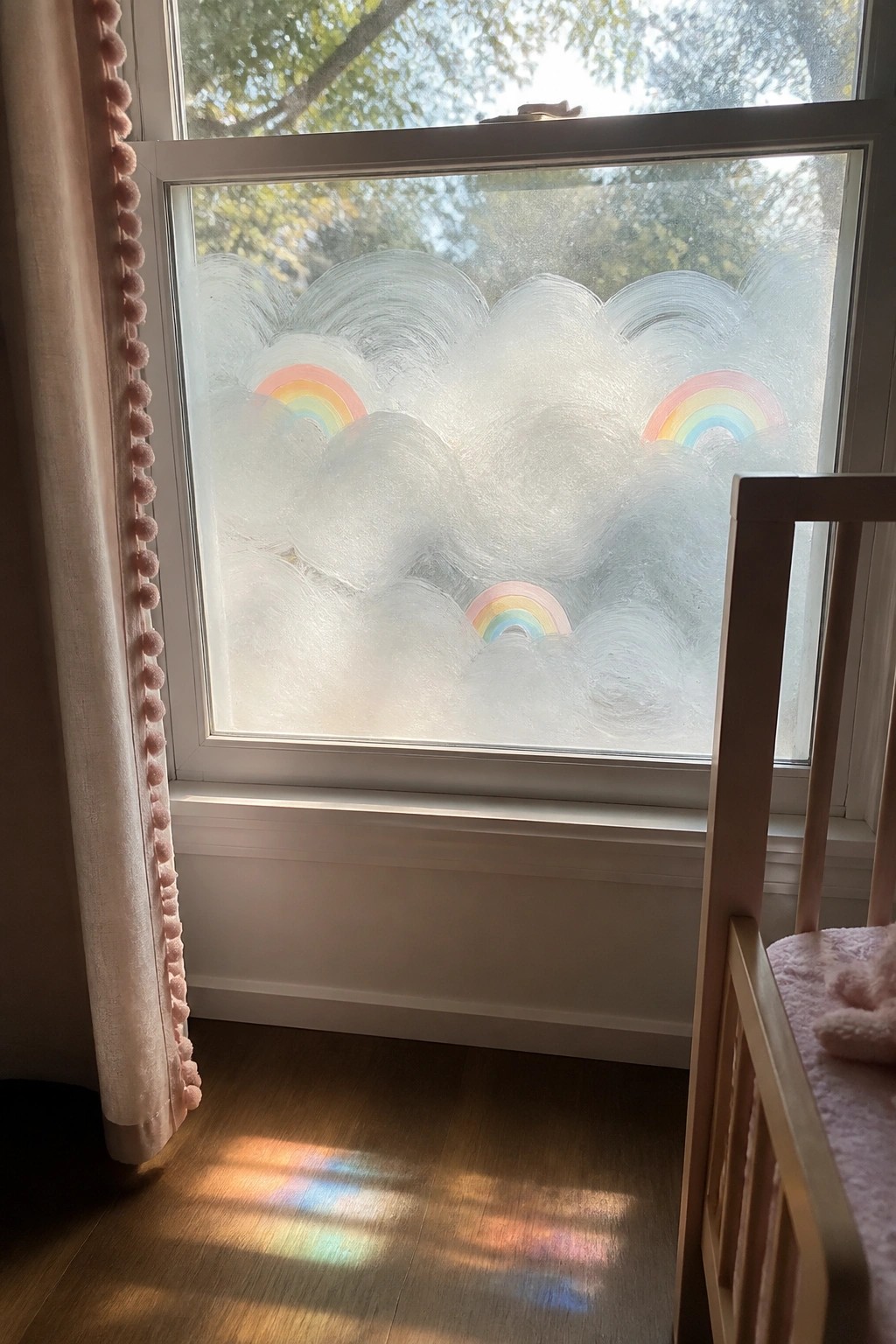 Window with cloud and rainbow decals casting colorful reflections on floor near pink curtain and crib