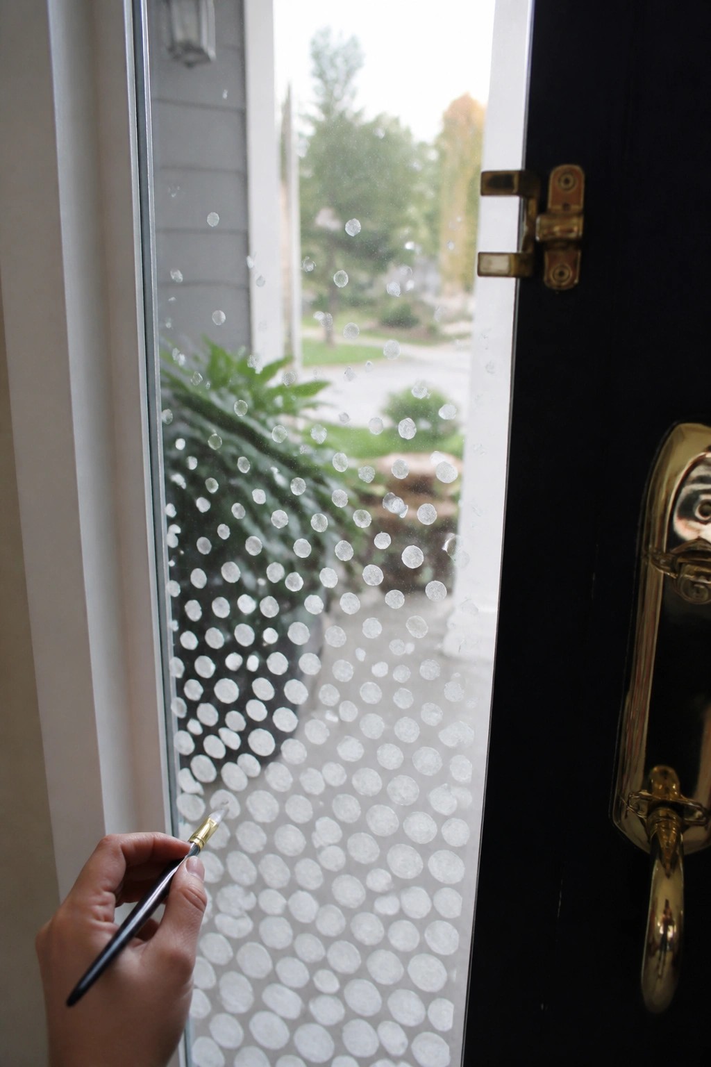 A hand holds a paintbrush applying white circular dots to the glass panel beside a black door, with the pattern growing denser lower down.