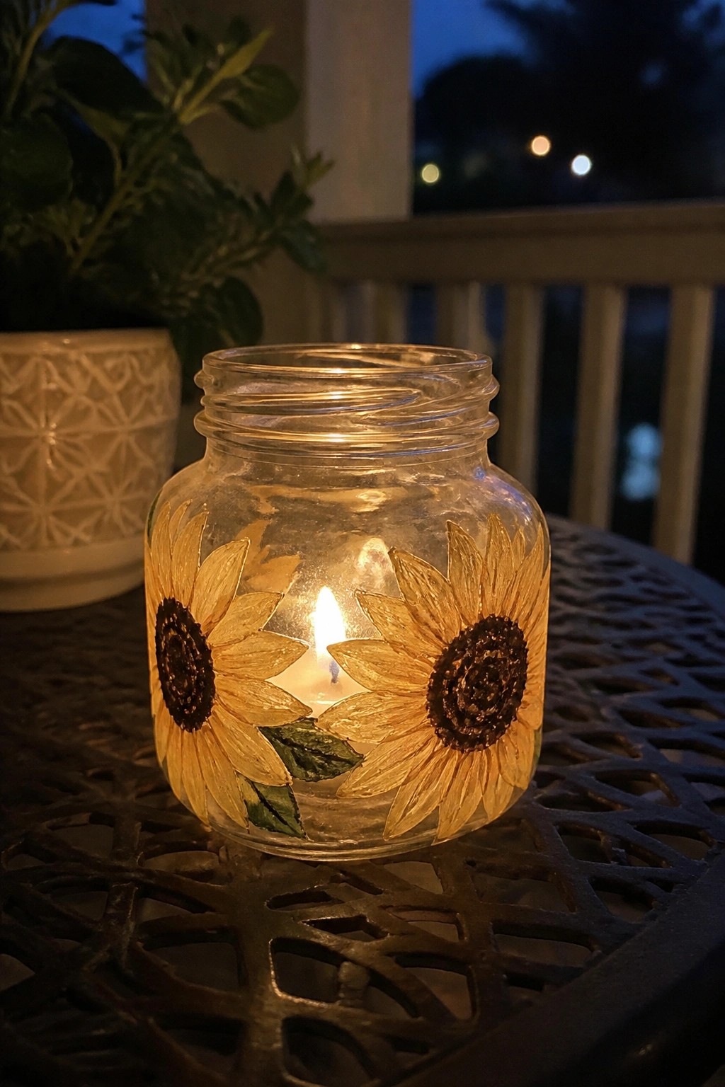 Lit candle glowing inside sunflower-painted glass jar on dark metal table at dusk.