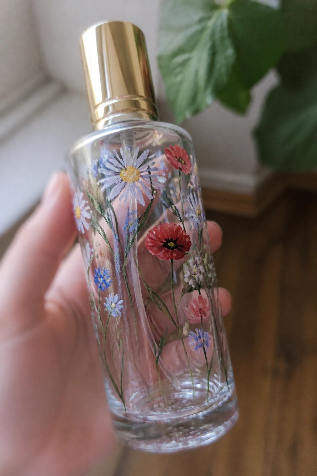 Hand holding clear glass bottle with gold cap and painted wildflowers