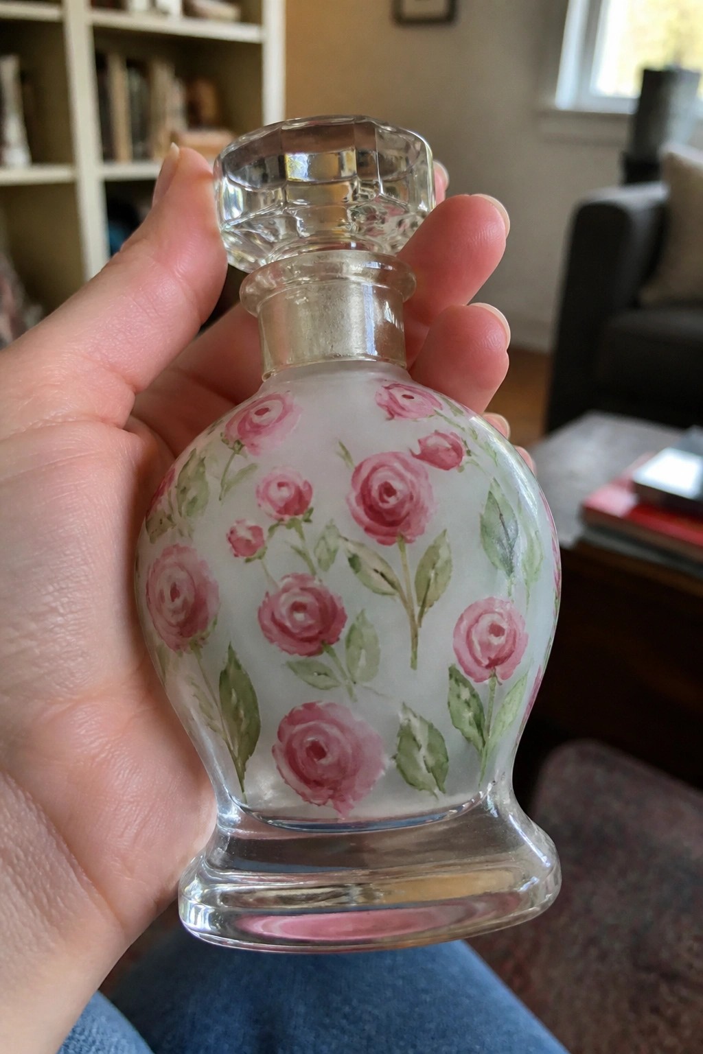 Hand holding a glass perfume bottle painted with pink roses and green leaves