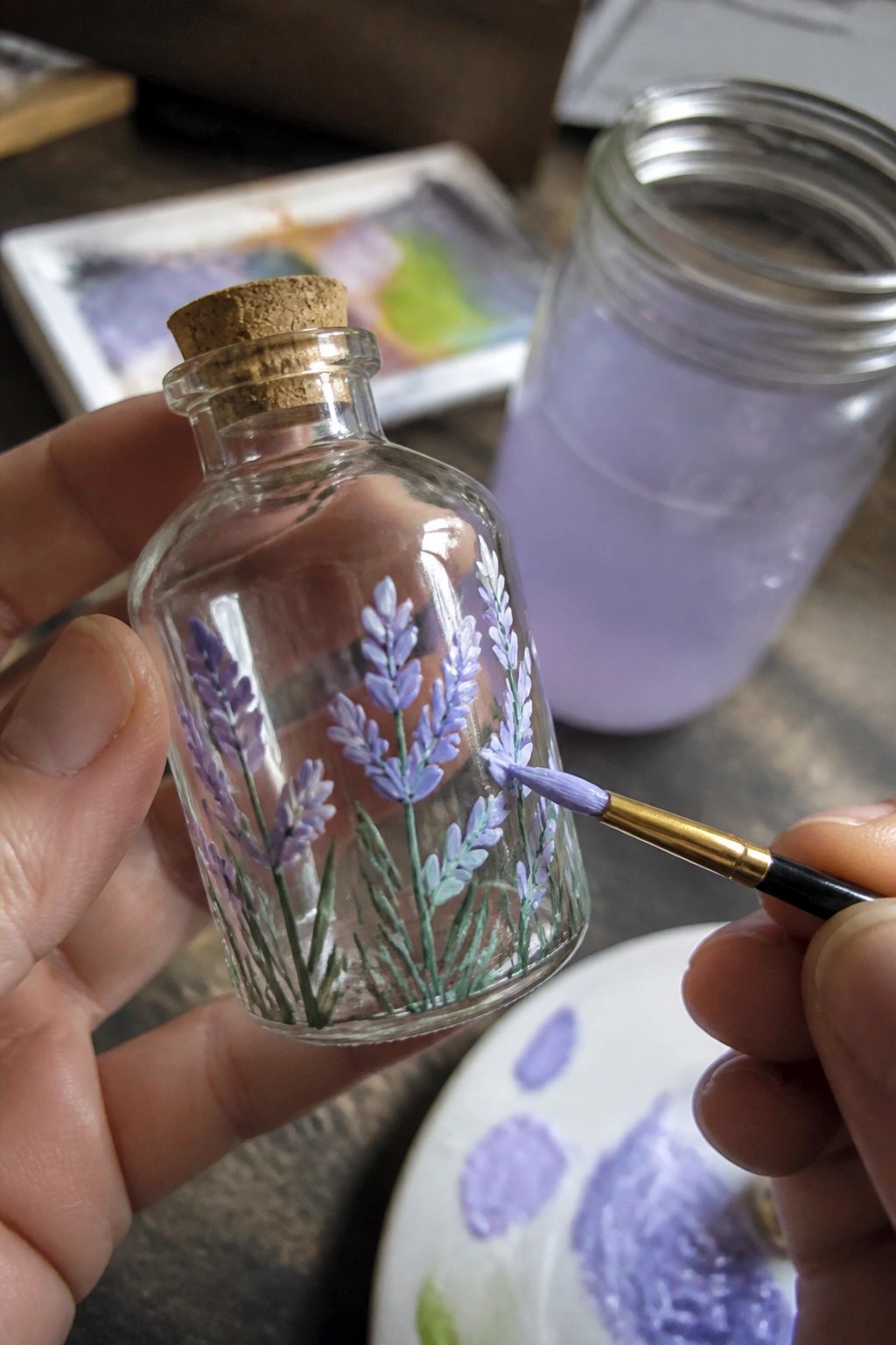Hands painting purple lavender flowers on a small corked glass bottle