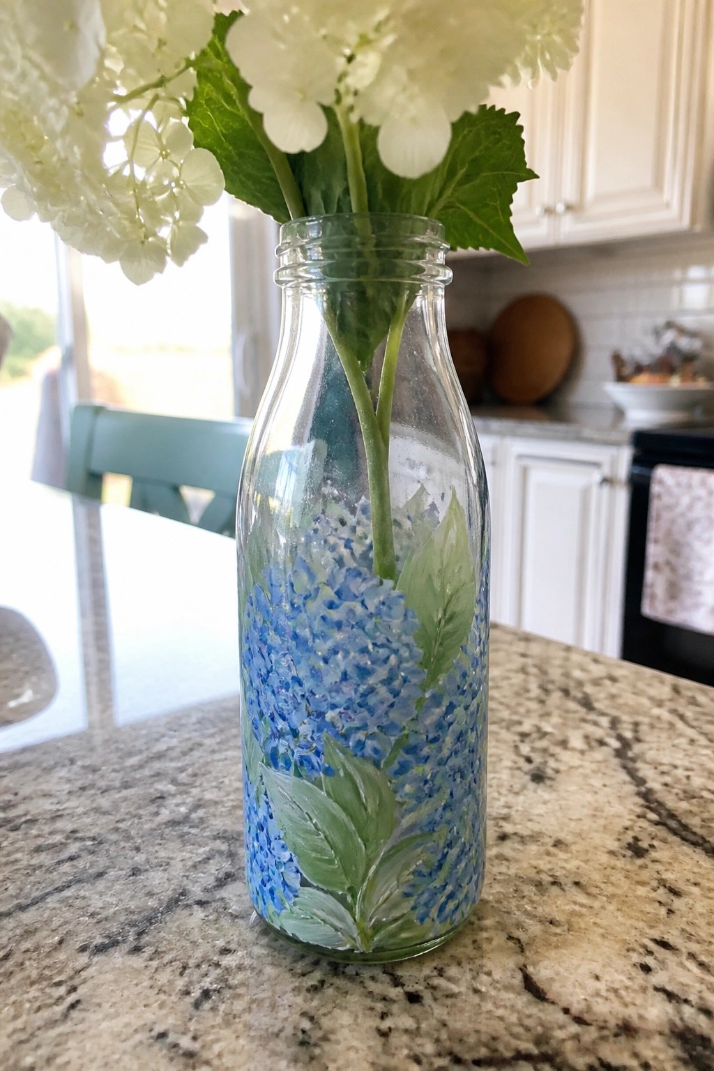 Glass milk bottle hand-painted with blue hydrangeas holding white flowers on kitchen counter