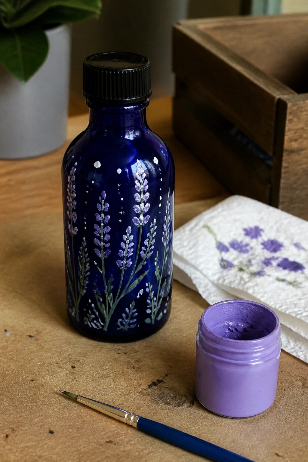 Blue bottle with hand-painted lavender flowers beside purple paint jar and brush on wood