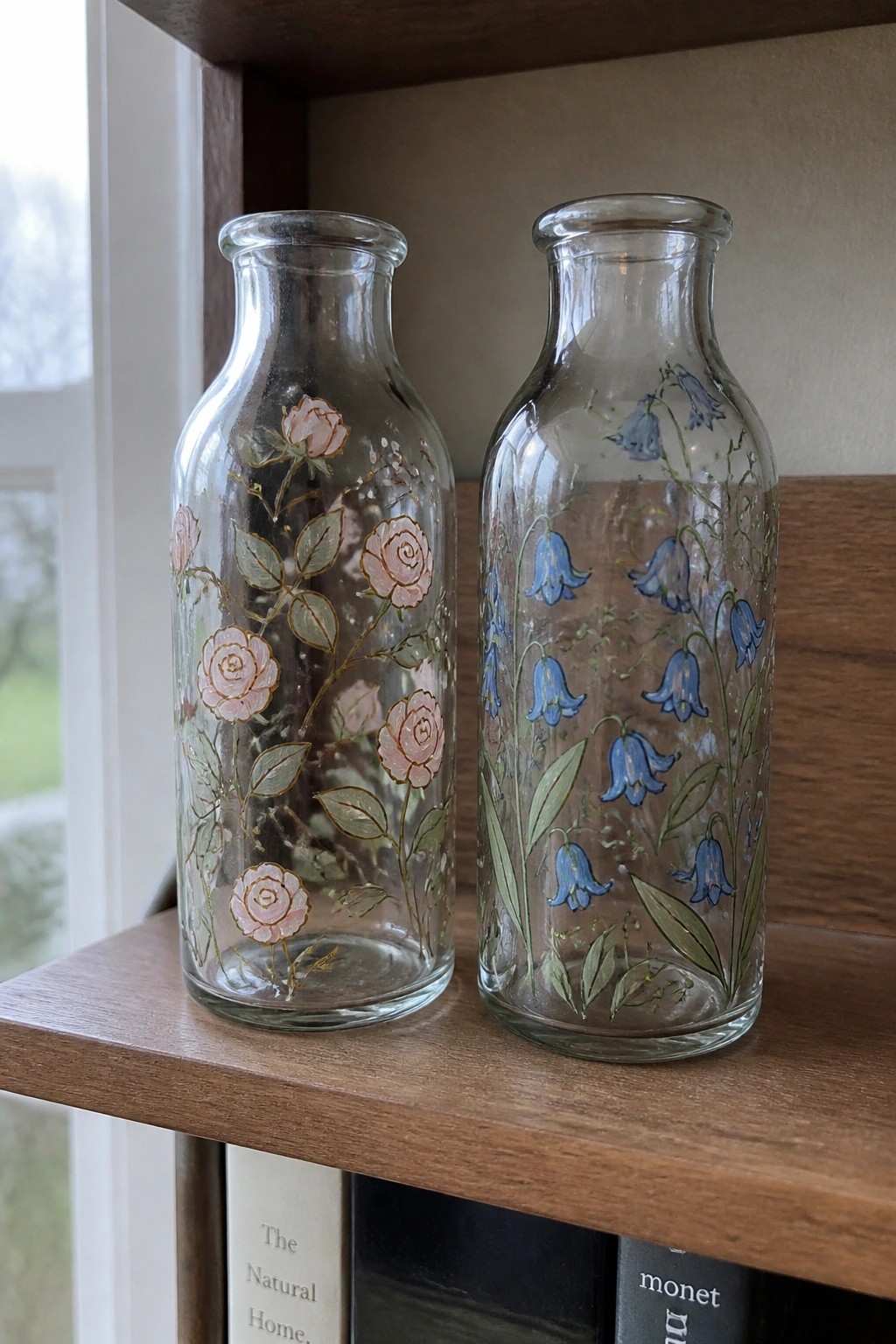 Two glass bottles painted with pink roses and bluebells on wooden shelf