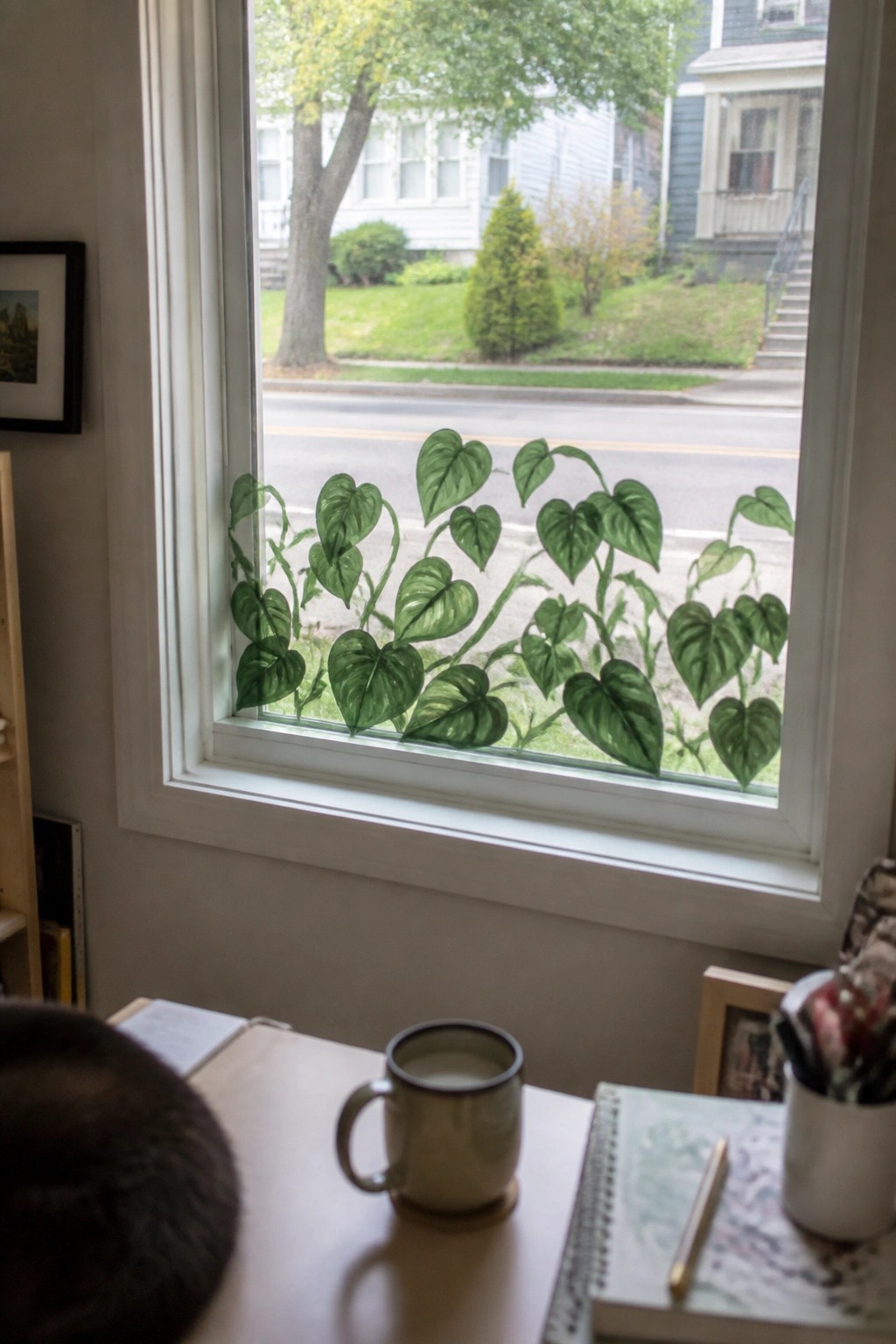 Window with green leafy vine decals, desk holding mug, notebooks and pen